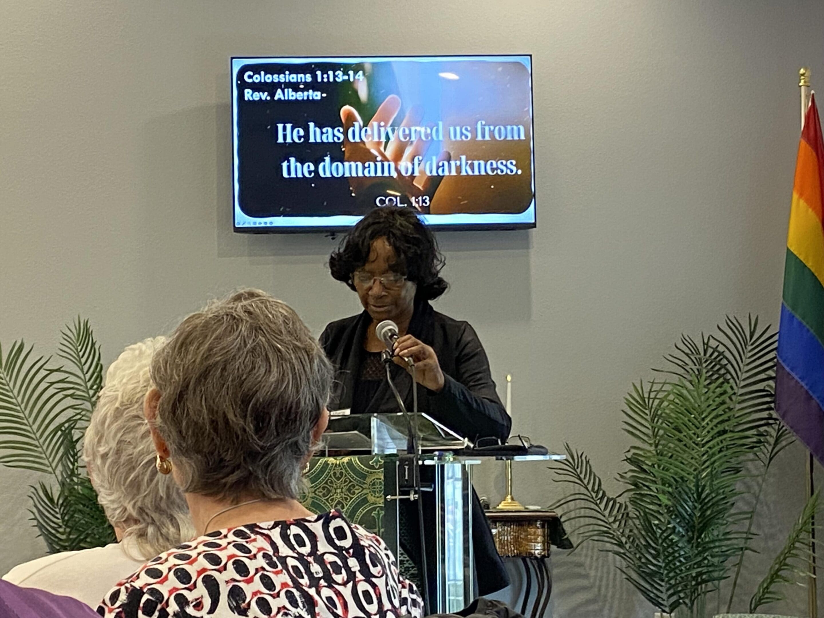 A speaker delivers a presentation with a screen showing text behind her.
