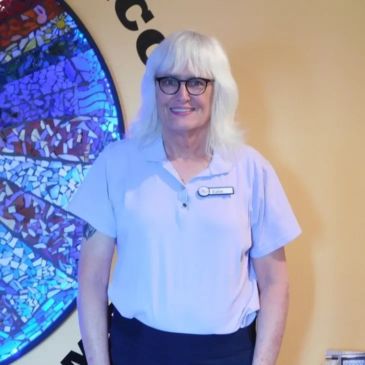 A woman with white hair and glasses smiling indoors.